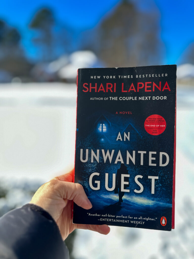 woman holds up An Unwanted Guest novel by Shari Lapena for best winter thrillers to read during a snow storm