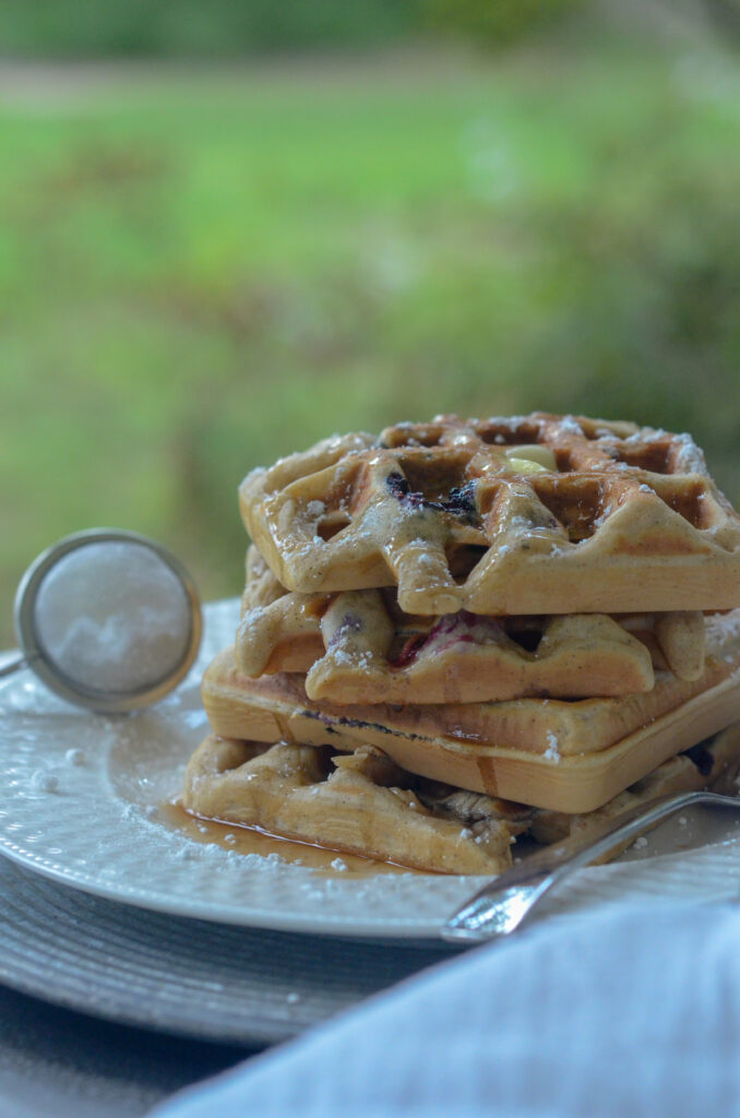 four blueberry waffles piled high on a plate with a powdered sugar dispenser to the side