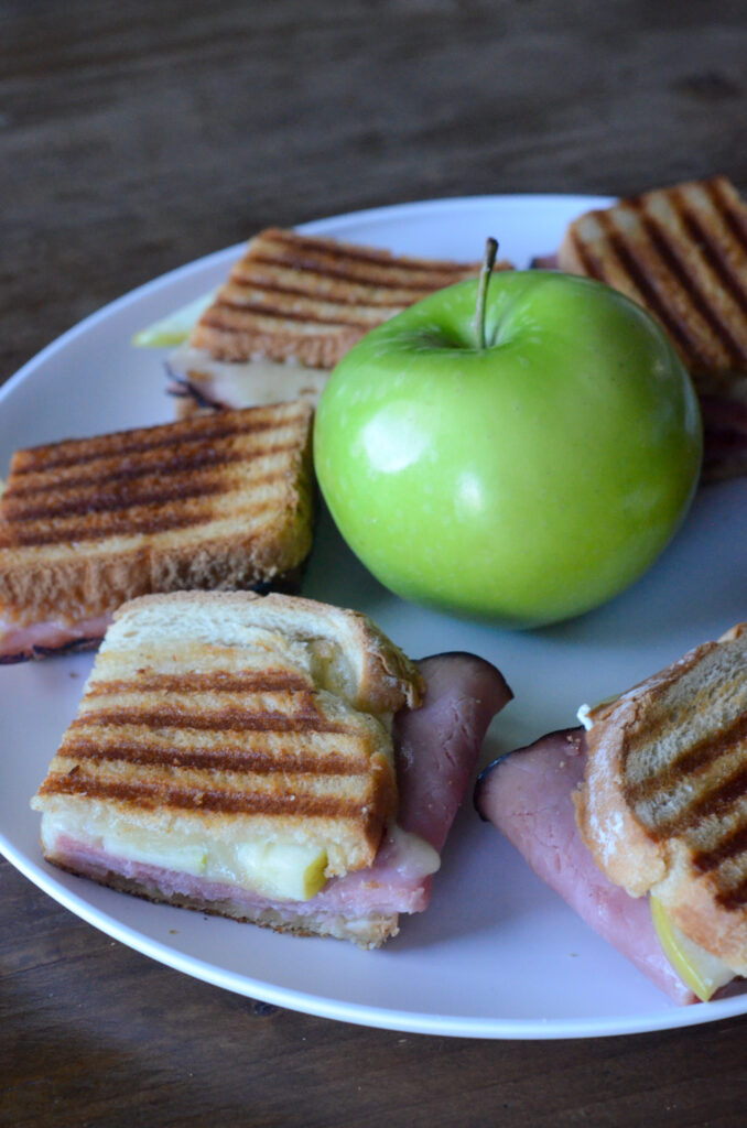 wedges of ham and cheese panini sandwich with a green apple in center of plate