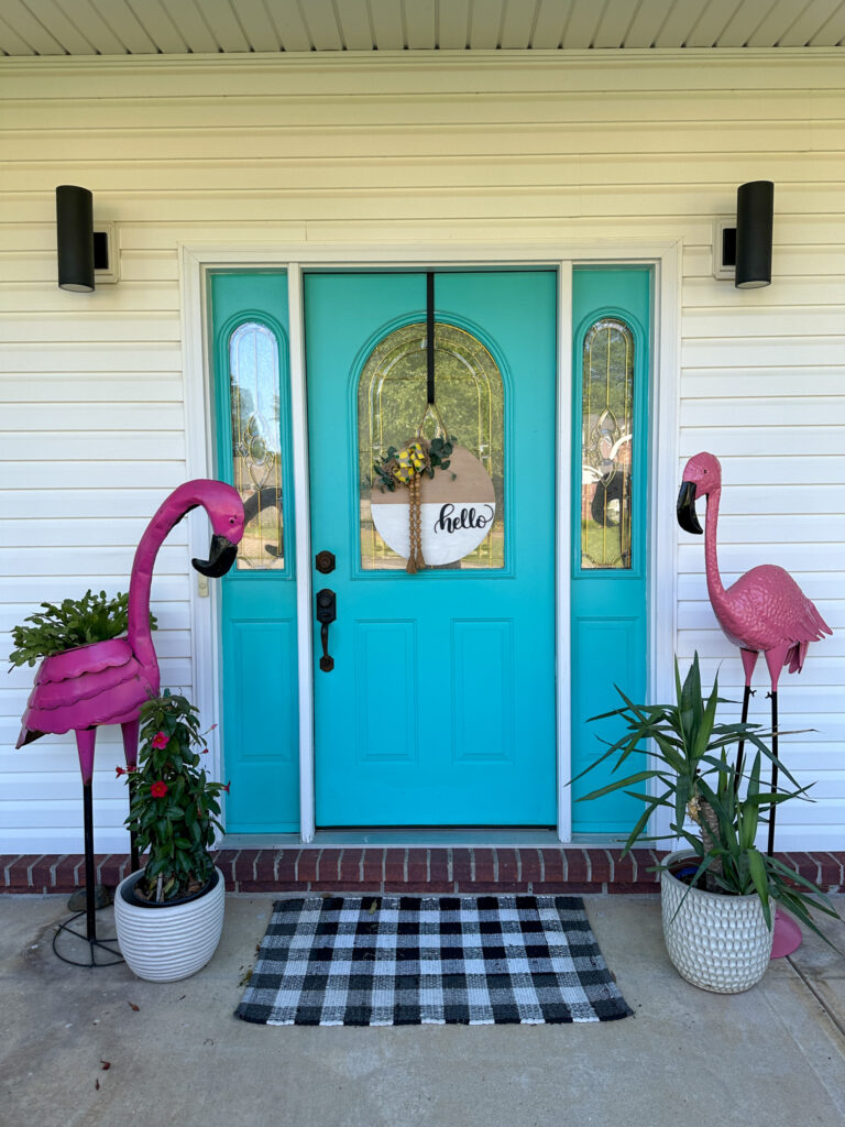plants and flamingos on a front porch with a turquoise front door