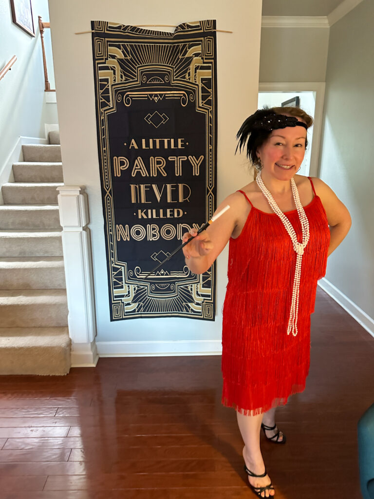 a woman at her 1920s murder mystery party in front of a "a little party never killed nobody" sign