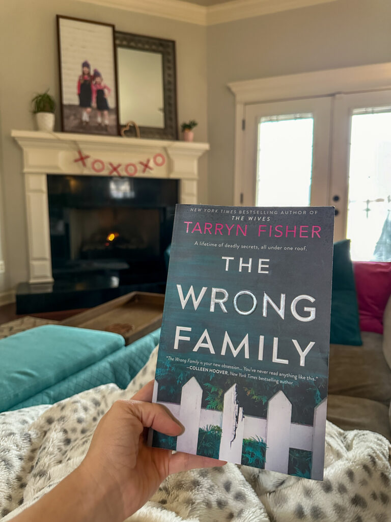 woman holds The Wrong Family novel in hand in front of fire