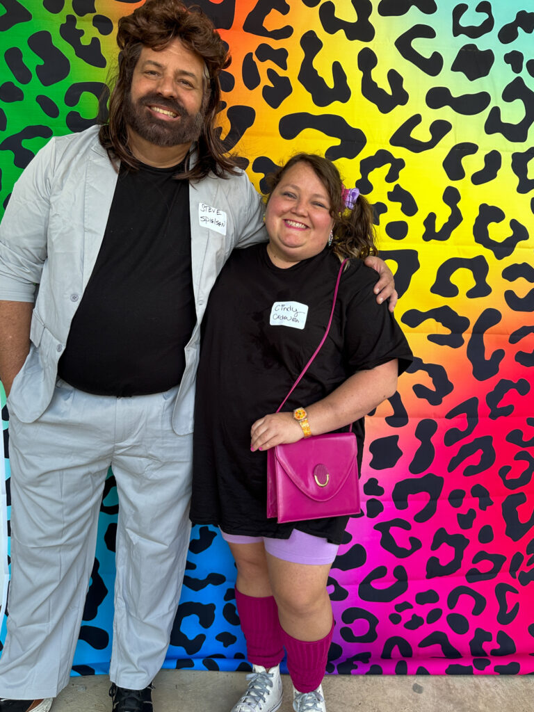 couple dressed in front of Lisa Frank style backdrop for 1980s murder mystery party