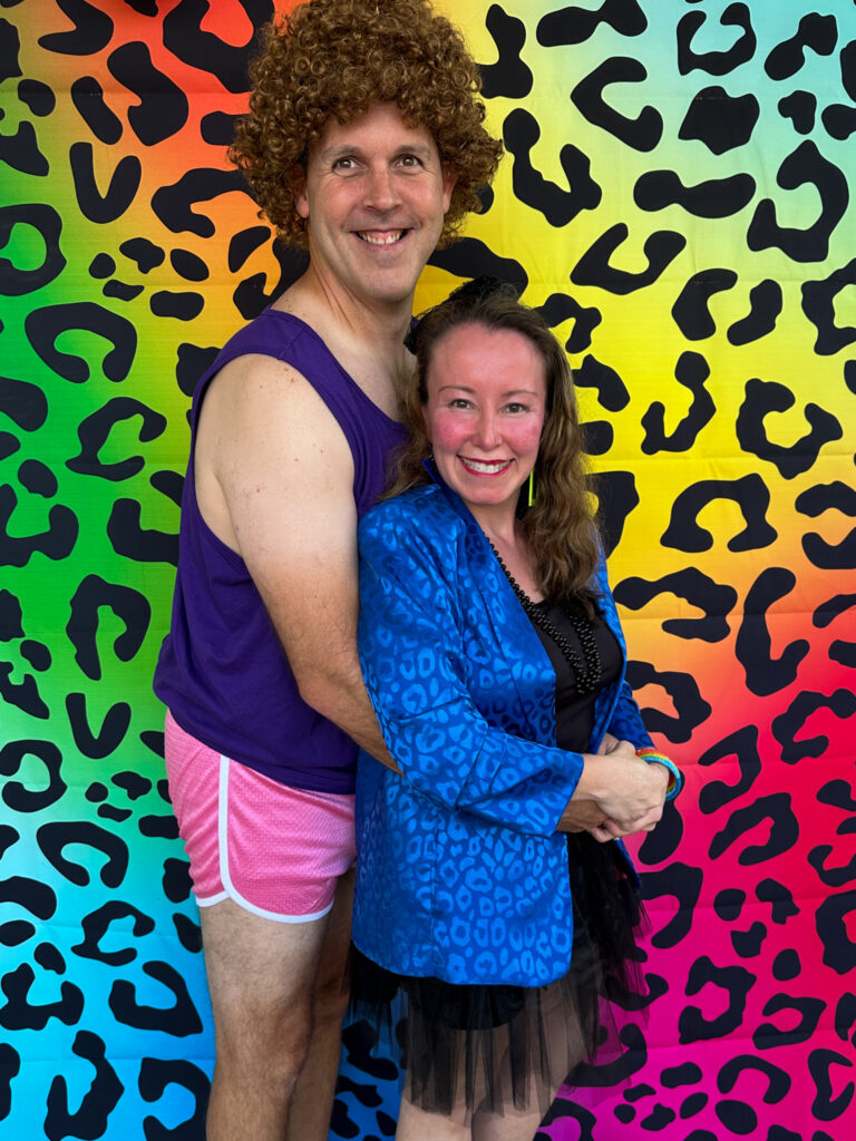 couple wearing men's 80s costume and women's 80s costume ideas in front of Lisa Frank style backdrop