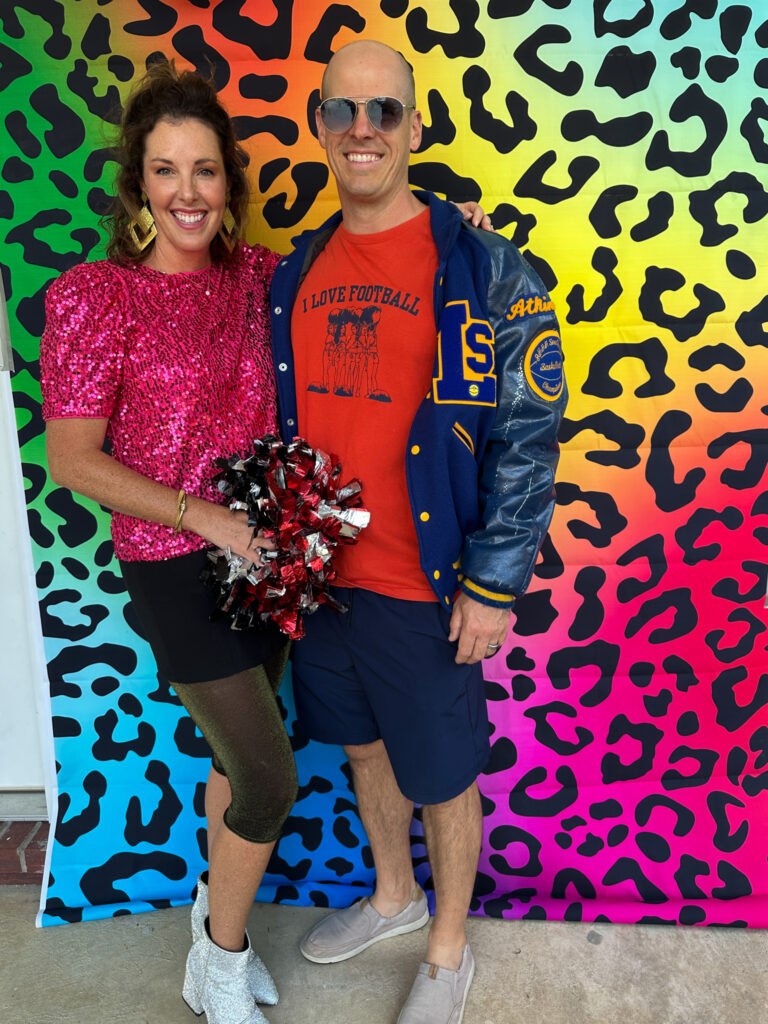 couple dressed in 80s costumes at 80s murder mystery party in front of animal print backdrop