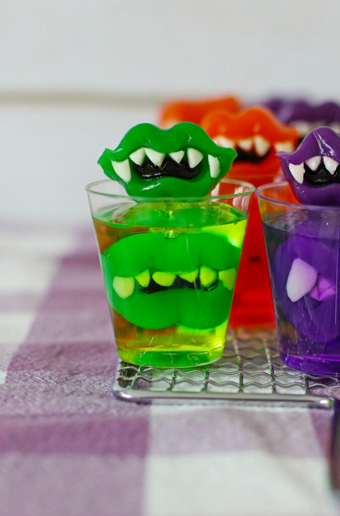 green, purple, and orange Jello shots with monster teeth for Halloween on metal tray and purple cloth