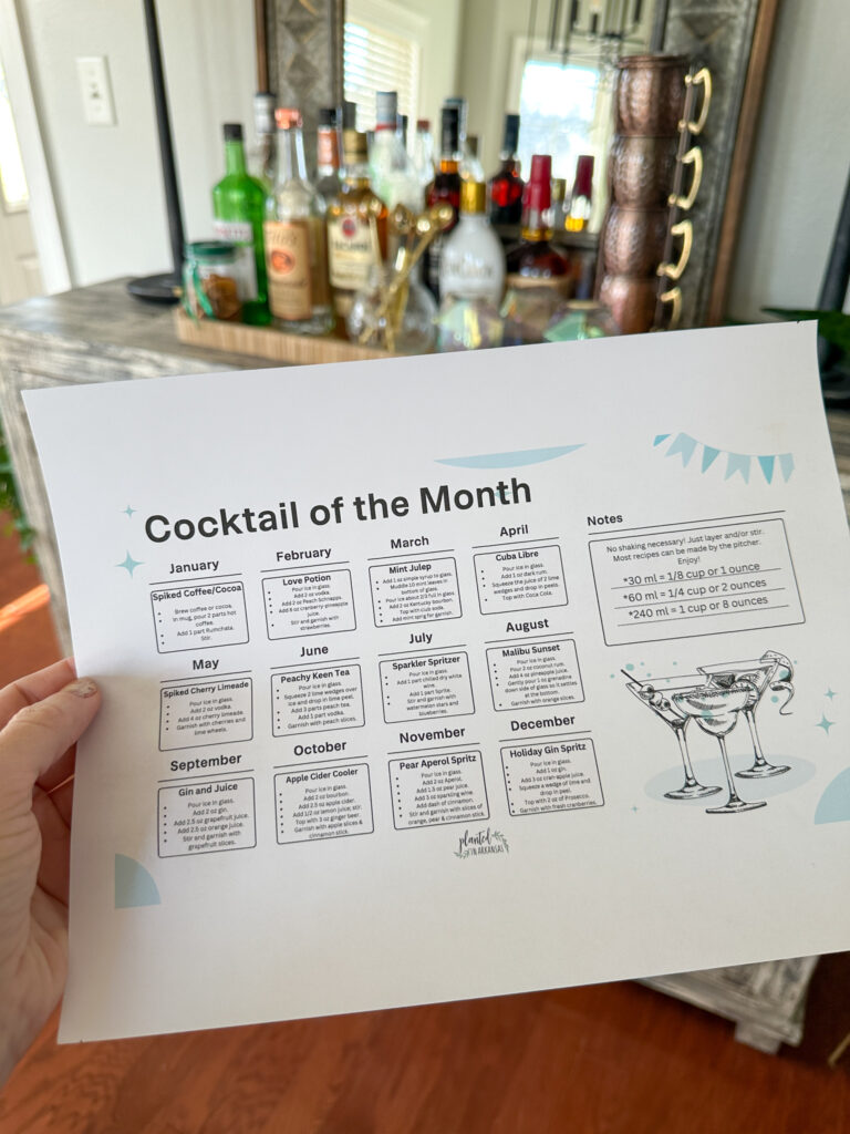 woman holds printable cocktail of the month calendar for planning in hand in front of bar tray