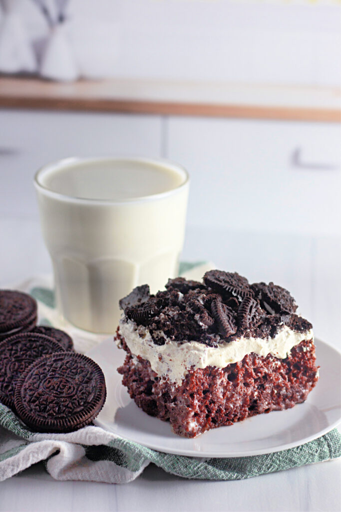 Oreo poke cake on white plate by glass of milk (cookies and cream poke cake)