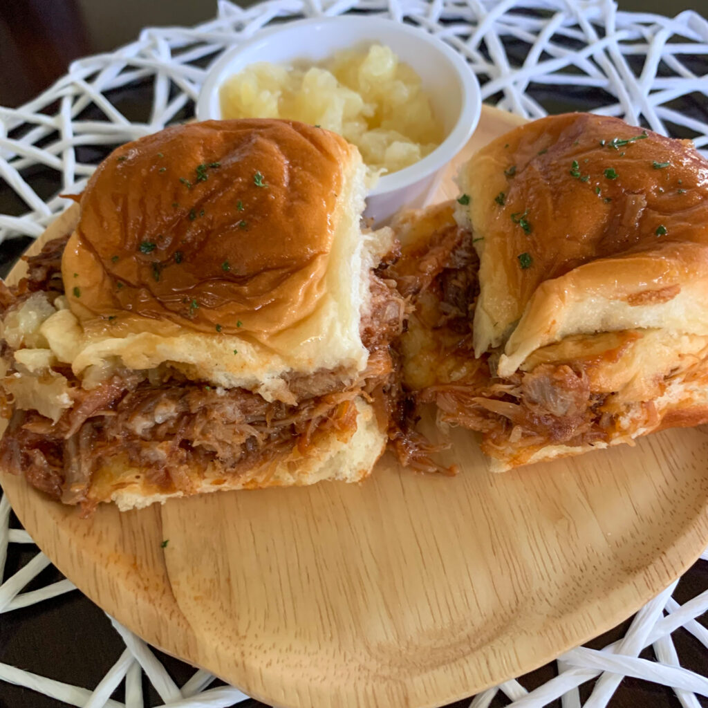 pulled pork pineapple sliders on Hawaiian rolls beside cup of pineapple on bamboo plate
