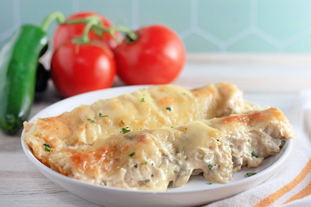 sour cream pulled pork enchiladas with green chiles on white plate with tomatoes and peppers at back