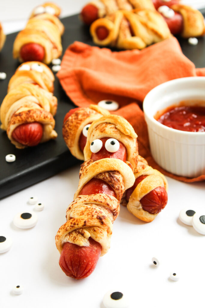 mummies in a blanket on white backdrop with dish of ketchup