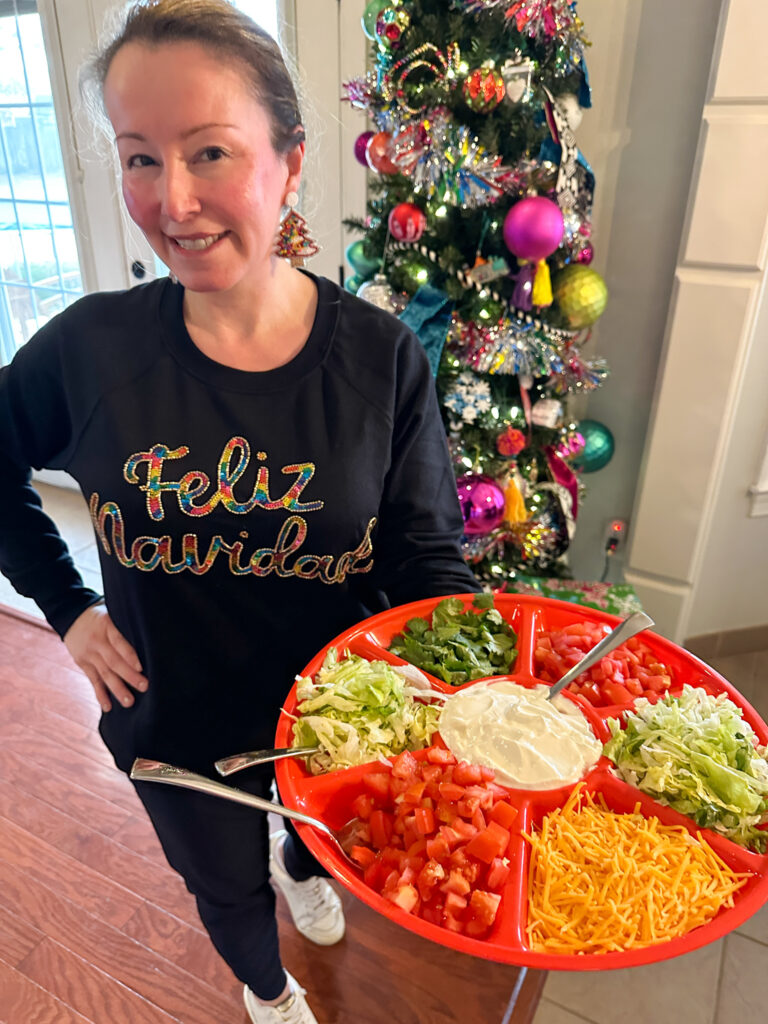 woman in Feliz Navidad sweatshirt holds tray of nacho toppings in front of fiesta style Christmas tree