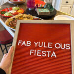 woman holds fab-yule-ous fiesta sign in front of Christmas dinner spread
