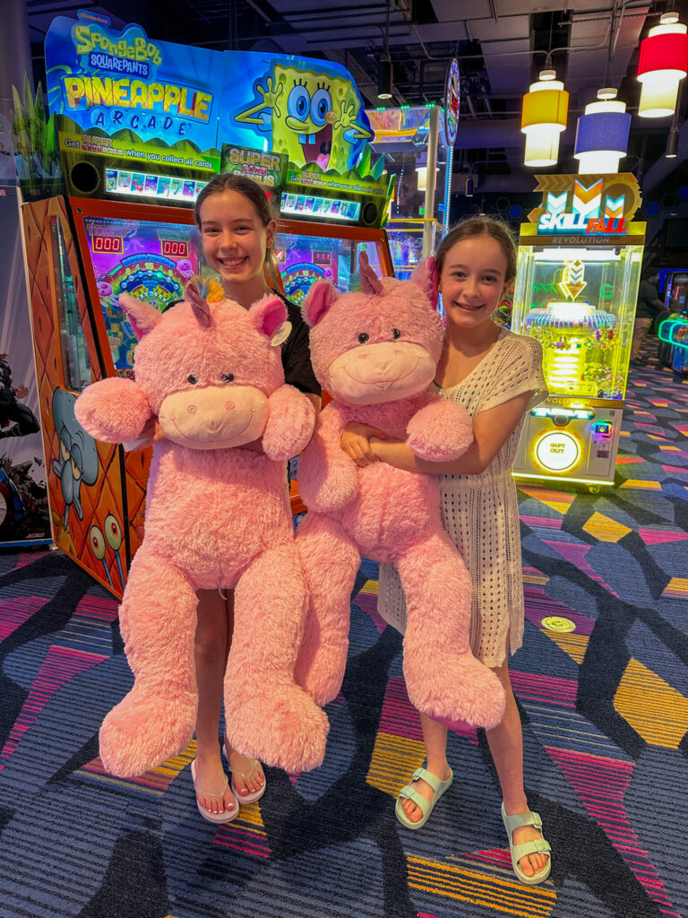 Arkansas bloggers, Harper and Norah, holding giant stuffed unicorns at Okana Resort and Waterpark arcade